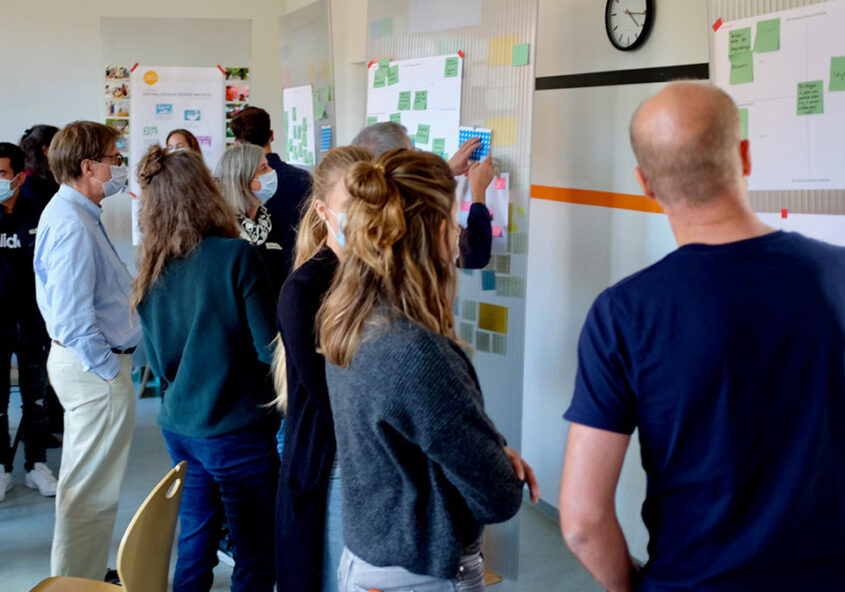 Gruppe von Menschen steht vor einer Wand mit vielen Zetteln und Notizen, einige Personen zeigen auf die Wand