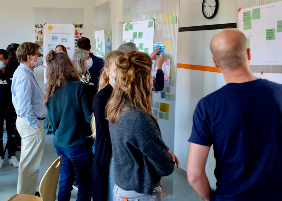 Home not Shelter_Ideen-Labor Gruppe von Menschen steht vor einer Wand mit vielen Zetteln und Notizen, einige Personen zeigen auf die Wand