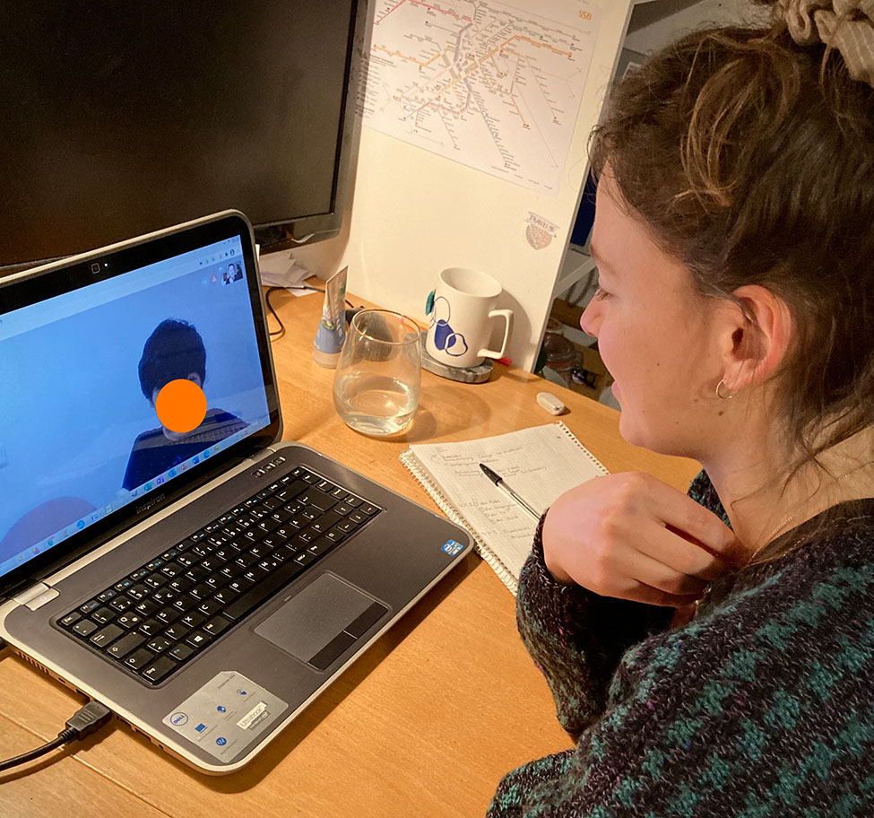 Home not Shelter_Lernpaten_Stuttgart Person sitzt an einem Schreibtisch vor einem Laptop mit einer Videokonferenz, auf dem Bildschirm ist eine Person mit orangefarbenem Kreis zum Anonymisieren vor dem Gesicht zu sehen