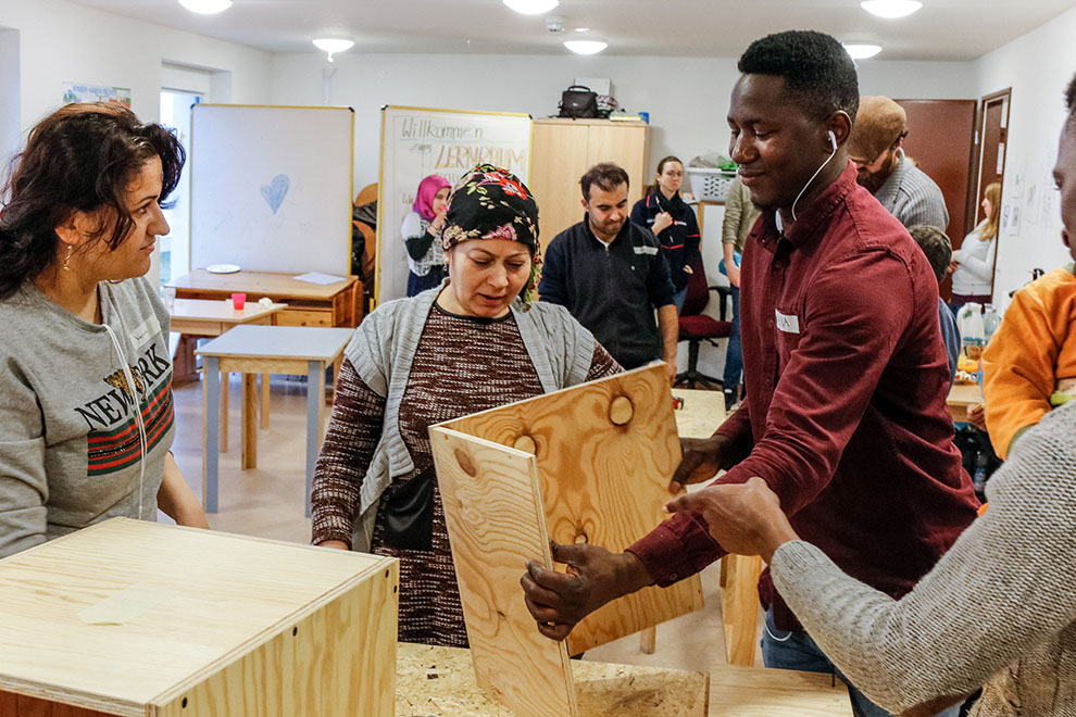 Home not Shelter_Lernraum Mehrere Personen in einem Raum greifen gemeinsam nach einem Holzobjekt, im Hintergrund Tische und ein Whiteboard mit Text