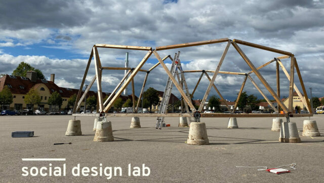 Home not Shelter_Sozialraumgestaltung_Cover Große offene Holzkonstruktion auf einem Parkplatz mit Betonfundamenten und Leiter bei bewölktem Himmel. Darunter im Bild das Textlogo des social design lab