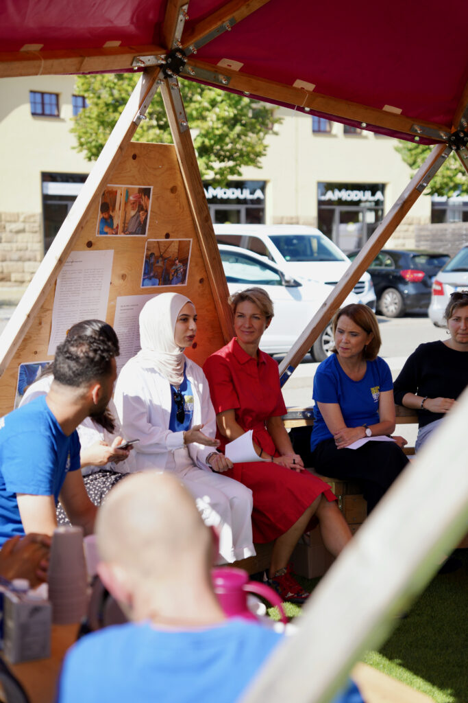 Gruppe von Menschen sitzt im Kreis unter einem roten Zelt, einige halten Notizb&uuml;cher, eine Frau mit wei&szlig;em Kopftuch spricht.