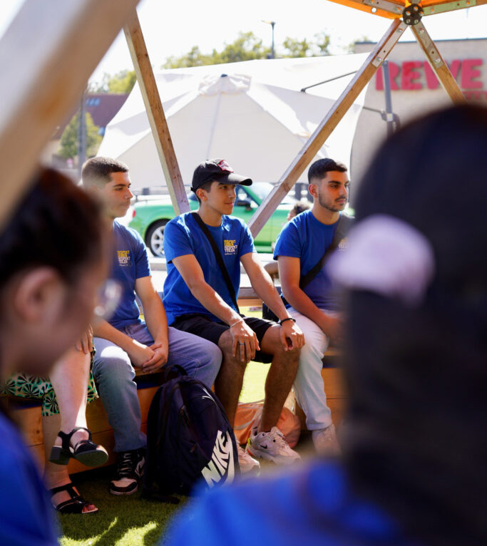 Home not Shelter_Transfer Mehrere Personen sitzen in einem Zelt mit Holzkonstruktion und grüner Bodenfläche, einige tragen blaue T-Shirts.