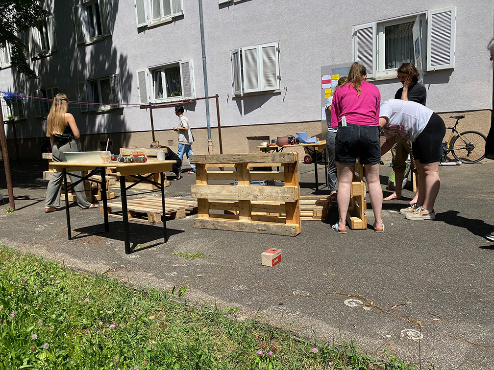 Home not Shelter_Werk-Stadt Mehrere Personen stehen und arbeiten an Tischen und einer aus Holzpaletten gebauten Konstruktion vor einem Wohnhaus mit weißen Fensterläden.