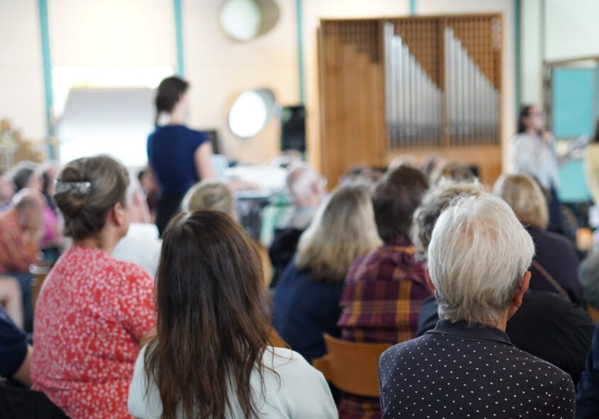 Es sind mehrere Menschen, die auf Stühlen sitzen von hinten zu sehen. Sie schauen alle nach vorne. Dort ist eine Orgel zu erkennen und daneben ein Aufbau mit Präsentation.
