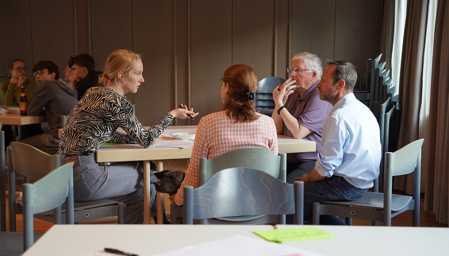 Roundtable_Werkstätten_Partizipation_Laim Fünf Personen sitzen an einem rechteckigen Tisch in einem hellen Raum. Eine Frau mit gemustertem Oberteil spricht gestikulierend mit den anderen, die aufmerksam zuhören. Die Gruppe wirkt konzentriert, einige haben die Hände am Kinn oder auf dem Tisch. Im Vordergrund ist eine leere Stuhllehne und ein Tisch mit einem Blatt Papier und einem leuchtend grünen Zettel zu sehen. Die Atmosphäre ist ruhig und sachlich.