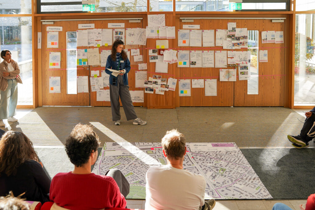 25102125_Projektschmiede_MO_HSS_Auswahl.bearbeitet_c.mayer_OK_11 Eine Gruppe von Menschen sitzt auf dem Boden und blickt auf einen großen Stadtplan, der auf dem Teppich ausgebreitet ist. Im Hintergrund steht eine Person vor einer Wand, die mit zahlreichen Blättern, Zeichnungen und Notizen bestückt ist. Die Wand besteht aus Holzpaneelen mit großen Fenstern und Glastüren. Die Szene wird von Tageslicht durchflutet.