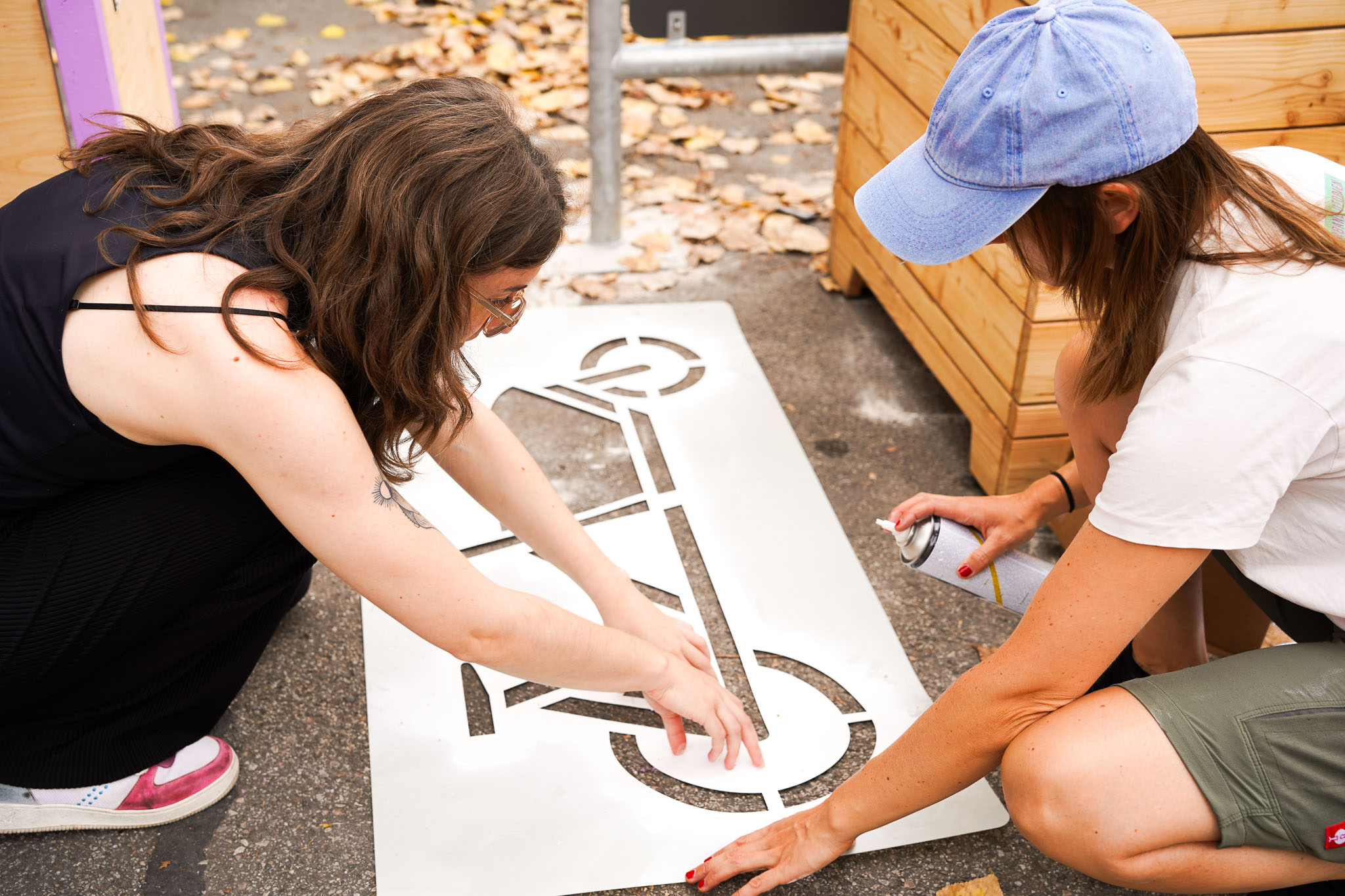 MOSAIQ Projektquartiere Fahrradreparaturstation Symbol Zwei Personen knien auf Asphalt und halten eine große Schablone mit dem Umriss eines Fahrrads. Die linke Person trägt eine Brille und hat lange, braune Haare, die rechte Person trägt eine blaue Baseballkappe und sprüht mit einer Spraydose weiße Farbe auf die Schablone. Im Hintergrund liegen herabgefallene Blätter und Holzelemente, die an Hochbeete erinnern. Die Szene wirkt wie eine dokumentarische Aufnahme einer gemeinschaftlichen Aktion im öffentlichen Raum und eignet sich als Cover eines Berichts.