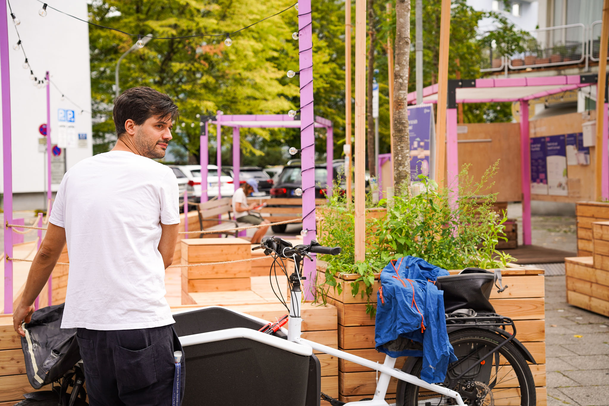 MOSAIQ Aufbauten Projektquartiere Im Vordergrund steht eine Person mit weißem T-Shirt und dunkler Hose neben einem Lastenfahrrad, das mit einer blauen Jacke und weiteren Gegenständen beladen ist. Die Szene spielt in einem städtischen Außenbereich mit Sitzbänken und Pflanzkästen aus Holz, die durch lilafarbene Streben und Dekorationen ergänzt werden. Im Hintergrund sind Bäume, Gebäude und eine Informationstafel zu erkennen. Die Gestaltung und die Farben der Umgebung wirken modern und einladend.