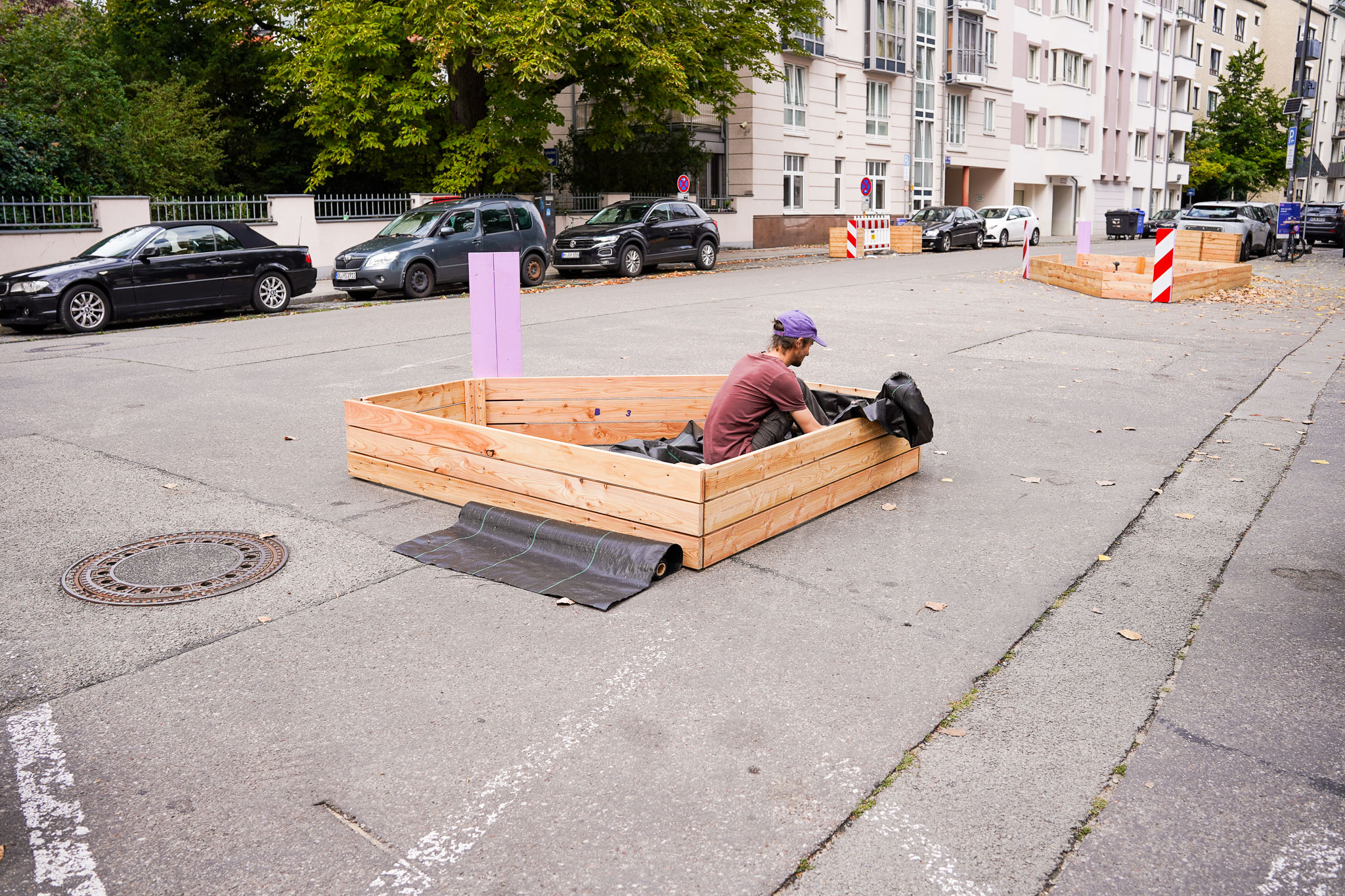 MOSAIQ Aufbauten Projektquartiere Schwabing West Querungshilfe Im Vordergrund befindet sich eine rechteckige Holzkonstruktion, die auf einer asphaltierten Fläche steht. Eine Person mit dunkler Kleidung und lila Mütze sitzt darin und arbeitet mit einem schwarzen Gegenstand. Die Konstruktion ist mit einer schwarzen Matte verbunden, die auf den Asphalt ausgelegt ist. Im Hintergrund stehen mehrere Autos am Straßenrand, daneben sind mehrstöckige Wohnhäuser mit Balkonen und große Bäume zu sehen. Absperrungen und ein lilafarbenes Schild markieren Bereiche der Straße. Die Szene vermittelt den Eindruck einer temporären Installation im urbanen Raum, passend zum Kontext eines Bericht-Covers.