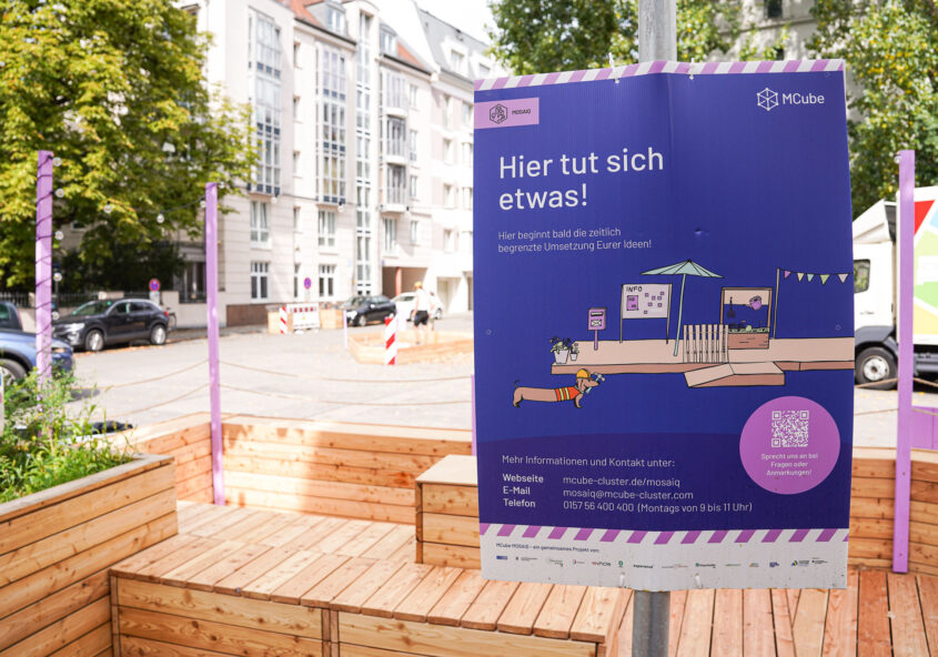 Im Vordergrund steht ein großes, lila Plakat mit weißem Text und einer stilisierten, farbigen Linienzeichnung einer Sitzlandschaft mit Sonnenschirm und Pflanzen. Das Plakat ist an einer Holzumrandung befestigt, die einen Bereich mit Sitzbänken und Pflanzkästen abgrenzt. Im Hintergrund sind mehrstöckige Wohnhäuser mit weißen Fassaden und zahlreiche Bäume zu sehen. Die Szene spielt im Freien, bei Tageslicht.