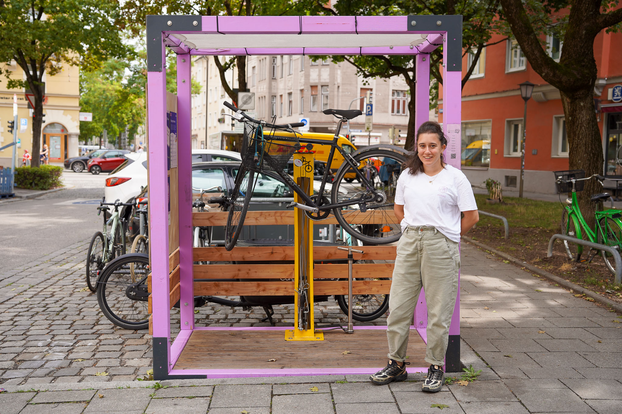 MOSAIQ Projektquartiere Fahrradreparaturstation Im Vordergrund steht eine junge Frau mit lockigem Haar, weißem T-Shirt und hellen Hosen, die entspannt lächelt. Sie befindet sich neben einer auffälligen, würfelförmigen Konstruktion mit lilafarbenen Rahmen und gelbem Fahrradständer, in dem ein Fahrrad befestigt ist. Im Hintergrund sind eine Holzbank, weitere Fahrräder und städtische Gebäude zu sehen. Die Szene spielt auf einem gepflasterten Platz mit Bäumen. Das Bild eignet sich als Cover eines Berichts, da es eine innovative städtische Fahrradstation und einen freundlichen, einladenden Moment zeigt.