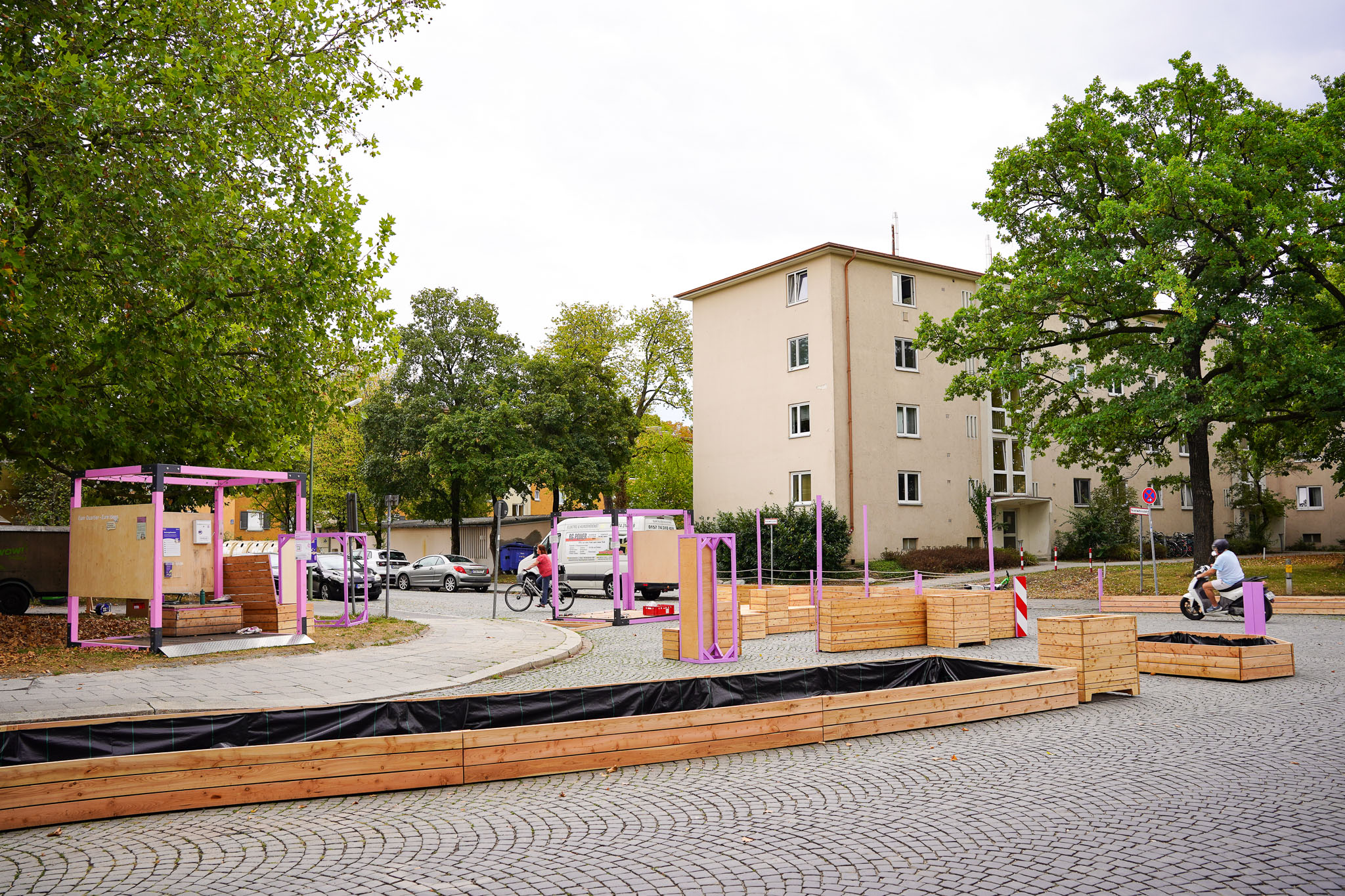 MOSAIQ Aufbauten Projektquartiere Moosach Kreuzung Im Mittelpunkt steht ein öffentlicher Platz mit mehreren rechteckigen Hochbeeten und Sitzgelegenheiten aus Holz, die von auffälligen, rosa lackierten Metallrahmen eingefasst sind. Im Hintergrund befinden sich mehrstöckige Wohnhäuser mit hellen Fassaden, umgeben von großen Laubbäumen. Auf dem Platz sind zudem Fahrräder und ein Motorroller abgestellt. Die Szene wirkt ruhig und geordnet, der Himmel ist bewölkt. Die Gestaltung und Möblierung deuten auf eine temporäre Umgestaltung oder ein Stadtentwicklungsprojekt hin.