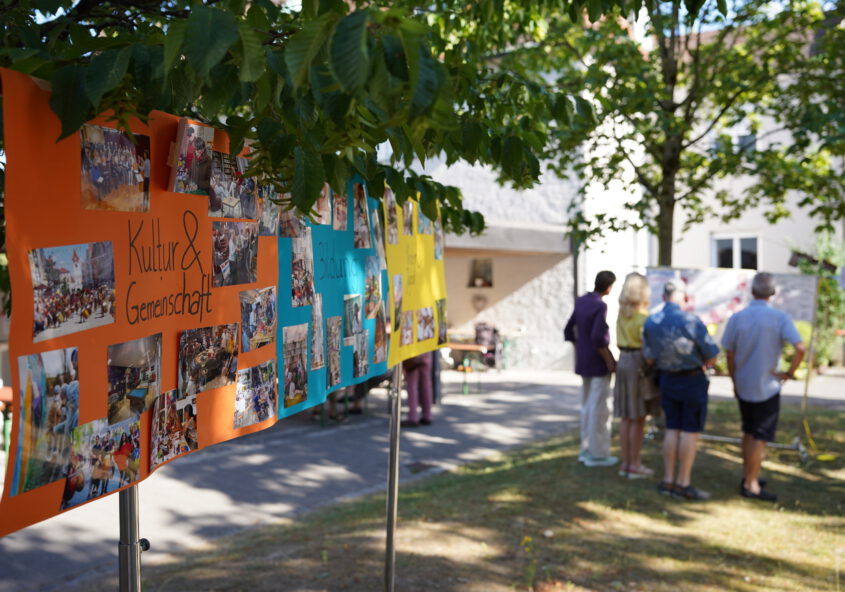 Zentral sind drei Poster in orange, blau und gelb zu sehen, die zwischen Bäumen aufgespannt sind. Die Poster bilden Themenbereiche ab, auf dem ersten ist "Kultur und Gemeinschaft" zu lesen. Dabei sind viele Abbildungen. Im Hintergrund ist eine Gruppe von Menschen zu sehen, die ein weiteres Poster, das zwischen anderen Bäumen augespannt ist, begutachten. Sie wirken konzentriert und stehen in der Gruppe beinander.