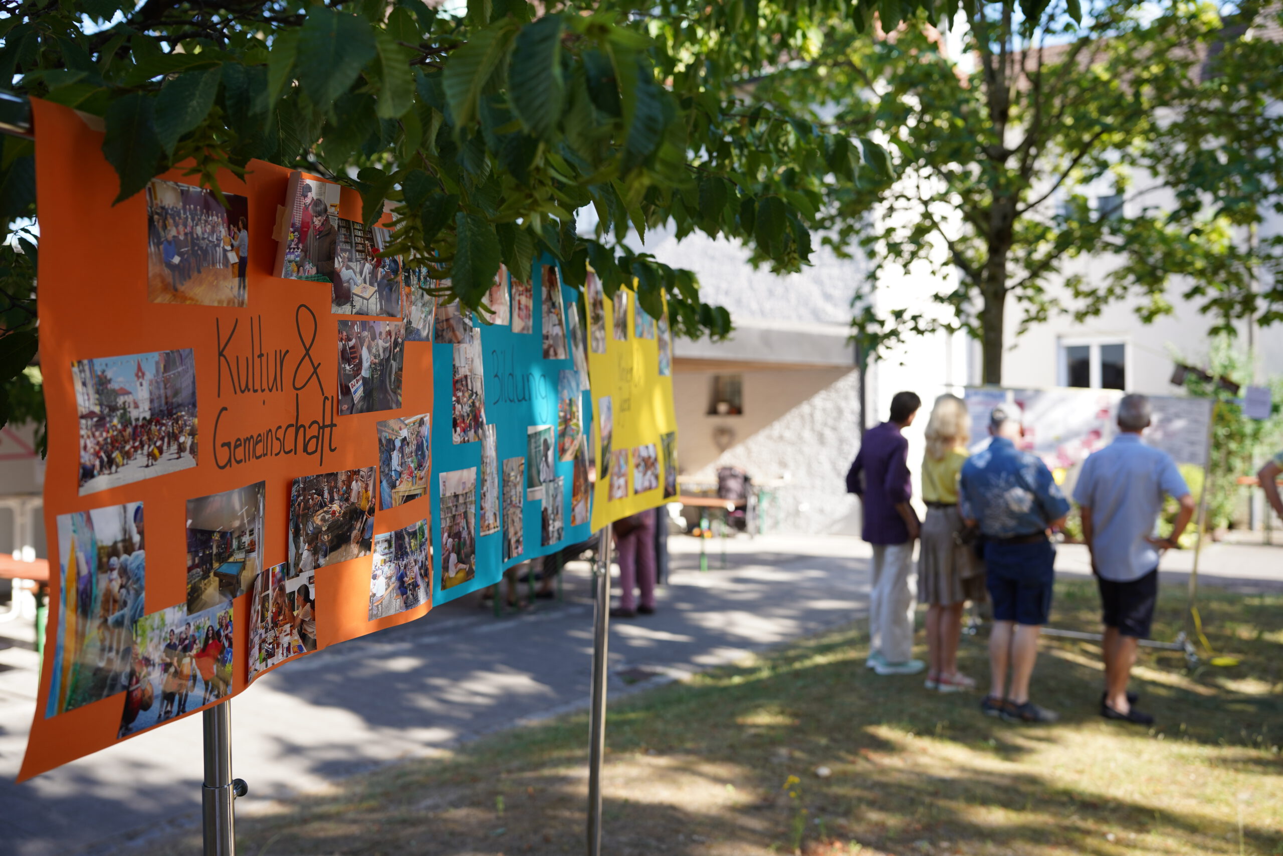 Zentral sind drei Poster in orange, blau und gelb zu sehen, die zwischen Bäumen aufgespannt sind. Die Poster bilden Themenbereiche ab, auf dem ersten ist "Kultur und Gemeinschaft" zu lesen. Dabei sind viele Abbildungen. Im Hintergrund ist eine Gruppe von Menschen zu sehen, die ein weiteres Poster, das zwischen anderen Bäumen augespannt ist, begutachten. Sie wirken konzentriert und stehen in der Gruppe beinander.